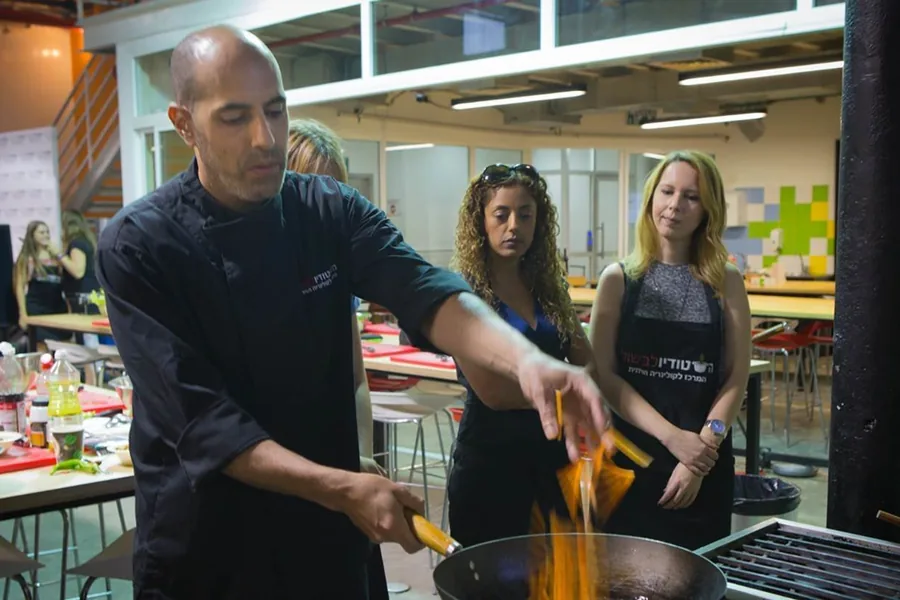 A chef pours oil into a pot, with participants working in the background